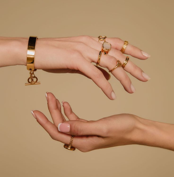 Two hands wearing gold jewelry against a beige background